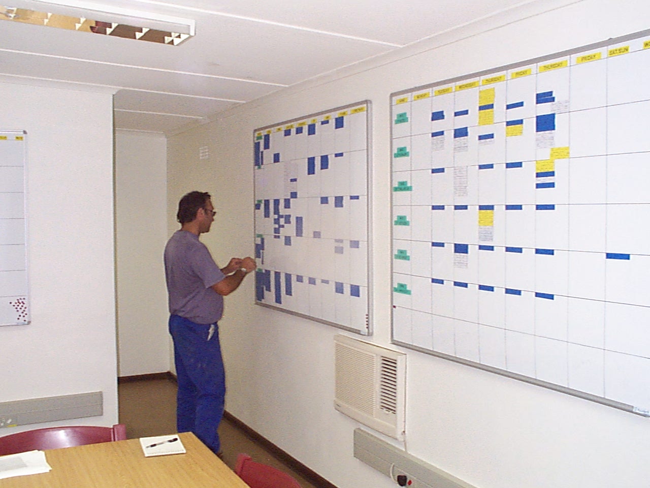 Maintenance planner standing at two wall-mounted whiteboard planning systems at the Koingnaas plant — a weekly planning-control board and a monthly look-ahead board, with colour-coded job cards.